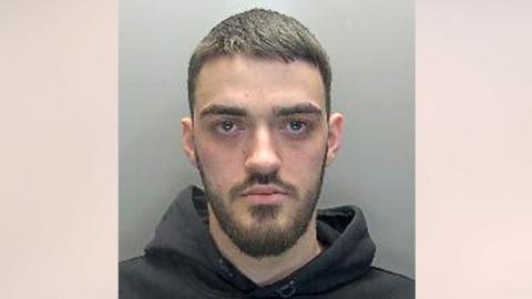 Lirim Shiqerukaj has short brown hair, and a moustache and beard. He is wearing a black hoodie and looking straight at the camera without any expression.