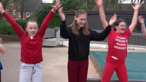 Three girls dressed in red outfits dancing on a school playground. They are standing in a line and have both of their arms up in the air in a Y shape. They are smiling.