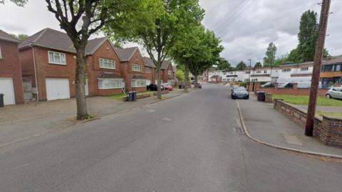 An image of a tree-lined road with houses on either side. There is a dark-coloured car parked in the distance.