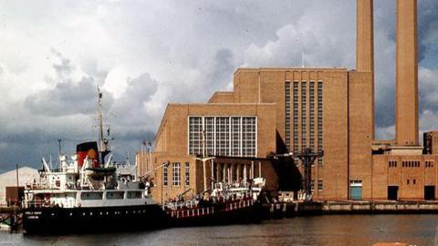 A waterfront brown brick building with two tall brown towers. A ship is moored alongside. 