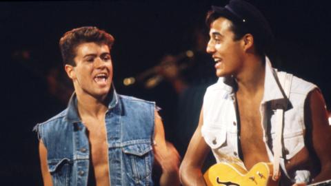 George Michael singing in a denim waistcoat and Andrew Ridgeley performing alongside him with a guitar while wearing a white waistcoast