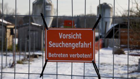 A red sign is strapped to locked gates. The sign reads VORSICHT SEUCHENGEFAHR! BETRETEN VERBOTEN! This translates to English as CAUTION DANGER OF DISEASE! ENTERING FORBIDDEN! There is snow in the yard behind the gates and two metal grain silos.