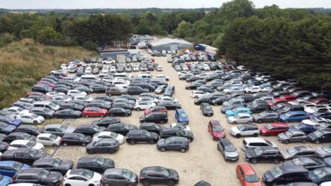 An aerial view of a large open-air car park. There are hundreds of cars lined up bumper to bumper, parked on gravel. There is a grey warehouse in the distance. The car park is surrounded by trees. 