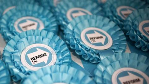 Light teal rosettes with the Reform UK logo - a white arrow point to the right with the word "Reform" in bold capitals - sits in the middle. The camera focusses on one rosette amongst rows neatly laid out on a white table. 