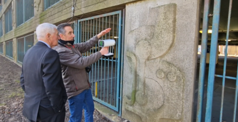 Two men stand beside a concrete panel, which is taller and wider than both of them. One man is holding a laser gun in front of him with his right hand and is pointing it at the concrete panel.