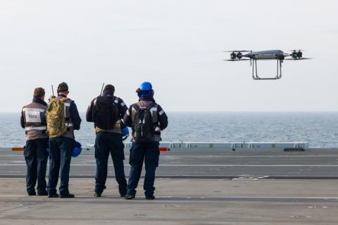 Image of the crew of four on the ship looking on at the drone as it hovers in the air