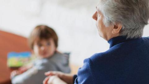 A grandmother sitting down with a young child in the background playing at a table.