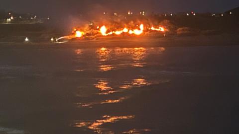 Flames in darkness of night on a beach, with flames reflected in the water.
