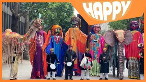 A parade of giant puppets, people playing drums and the Happy logo
