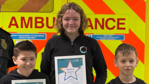 Three children standing in a row in front of a marked fluorescent ambulance. They are each looking at the camera and smiling and holding a certificate.