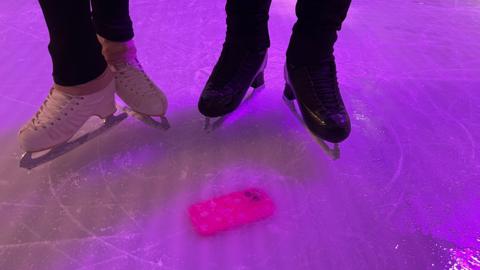 Bright pink phone case in ice. Standing next to it are the boots of two ice skaters. One set of white skates, to the left, and one set of black skates, to the right.