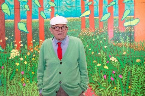 Hockney is wearing a hat, glasses, a green cardigan and a blue shirt and red tie. 