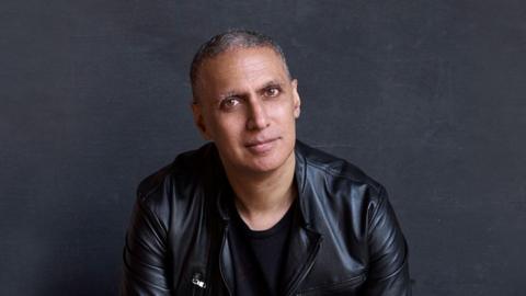 Nitin Sawhney wearing a black t-shirt and a leather jacket smiles for a professional photograph against a black background. 