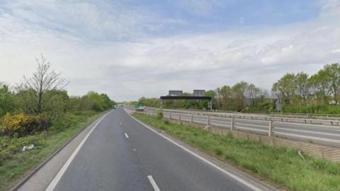 The left carriageways of a dual carriageway. The sky is blue but cloudy. There is green grass either side of the carriageway. No cars are visible.