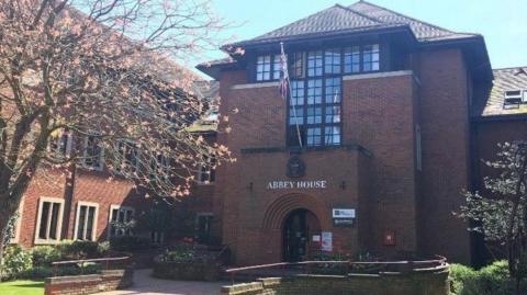 An exterior view of the Vale of White Horse District Council offices at Abbey House. It is a dark red brick building with black framed windows. The entry way to the building has an archway with a sign reading Abbey House above it and a Union Jack flag.