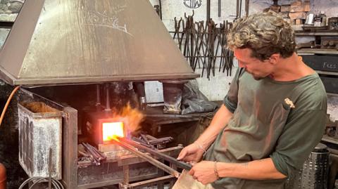 A man, wearing a green t-shirt and beige shorts, holding metal poles which are placed inside a burning furnace. He is in a workshop, surrounded by lots of metal items on shelves. A red gas cylinder is visible to the left of the image, as is an electrical fan.