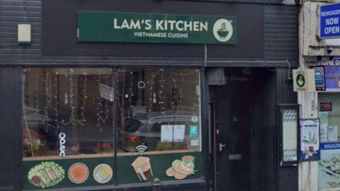 Lam's Kitchen Vietnamese restaurant on Otley Road in Leeds with a black wooden exterior, a green and white sign, and pictures of food with lights and an 'open' sign in the window.