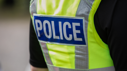 The back of a police officer, they are wearing a hi-viz vest that says police and a black top underneath