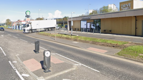 A crossing island in the middle of a road. One side of the road has a Starbucks drive thru and a sign for Burger King in the distance. A lorry waits to join the main road