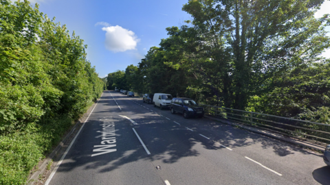 An A-road with cars parked on one side, with trees lining both sides of the road