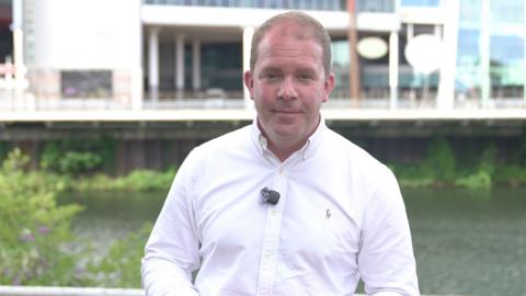 A man in a white shirt is looking directly into the camera. The background is the River Taff and an out of focus shot of the side of the Principality Stadium