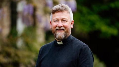 A man with a beard wearing a cassock with a clerical collar. He is smiling and standing outside in a garden area, although its details have been blurred by a soft focus to put emphasis on the man.