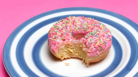 A ring donut with pink frosting and hundreds and thousands and a bite taken out of it, on a plate