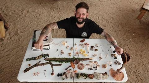 Andy Knott, who has a dark hair, a thick brown beard, and tattooed patterns down his arms, poses by a table which has a variety of different fungi laid out.