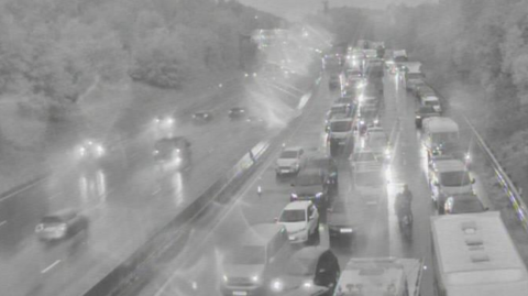 A traffic camera is showing a motorway which has four lanes on either side of the road. Traffic on the right is standstill in the black and white image.  