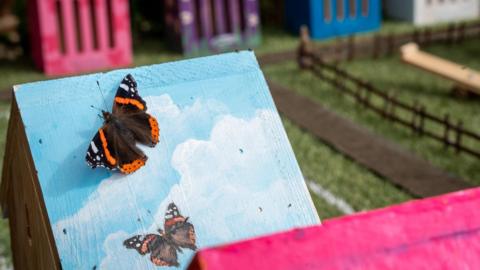 A black and orange butterfly sits on the slanted roof of a miniature house designed for butterflies, which is painted blue and white like the sky.