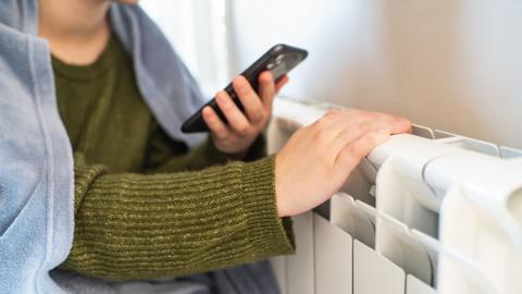 A person wearing a green knitted top and a light blue blanket around their shoulders holds a black smartphone while touching a white radiator. The scene is indoors with neutral-coloured walls.