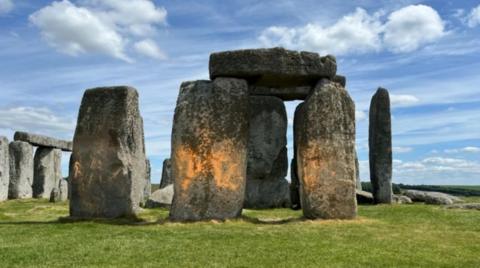 Image of Stonehenge with red paint marking two of the stones in the centre of the circle - surrounded by green grass and blue skies with fluffy clouds.