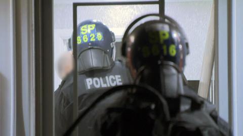 Police officers wearing protective helmets and tactical gear move through a doorway inside a building; “POLICE” and identification markings are visible on their helmets and backs.