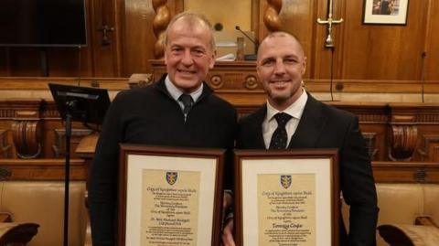 Neil Hudgell and Tommy Coyle receiving their awards at Hull's Guildhall. Both men are wearing dark jackets and a shirt and tie. They are standing in the council chambers holding the awards in front of them.