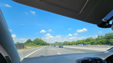 A shot taken from the passenger seat of a car looking at the motorway ahead. The sun is shining there are a few cars ahead and you can see cars over the central reservation going in the opposite direction. The sky is blue and punctuated by white fluffy clouds. Trees line the road on either side.