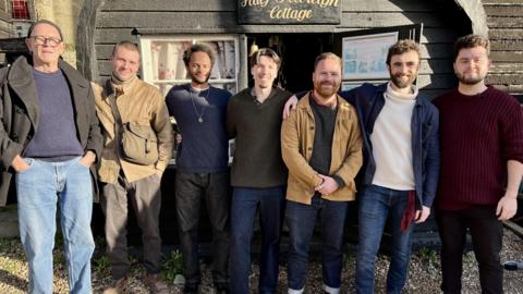 A group of seven men stood side by side with their arms around each other in front of a fishing hut.
