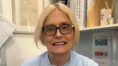Liz Wynn has blonde hair and is wearing a pale jumper and black glasses. She is sitting in a small office with various items on a shelf next to her.