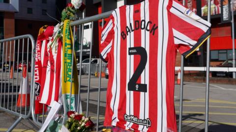 A red and white football shirt with Baldock written on the back, hanging on some grey metal fencing