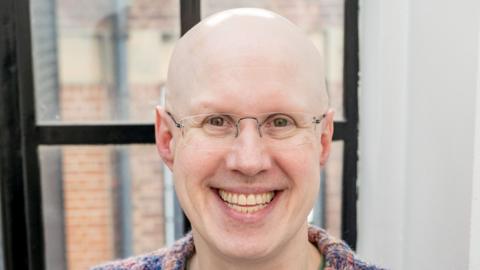 Matt Lucas, wearing glasses, smiles at camera in colourful jumper