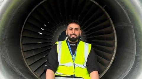 Faizan Mujtaba, with a beard, is smiling. He is sitting in the centre of the photo wearing a high-vis jacket and a lanyard.