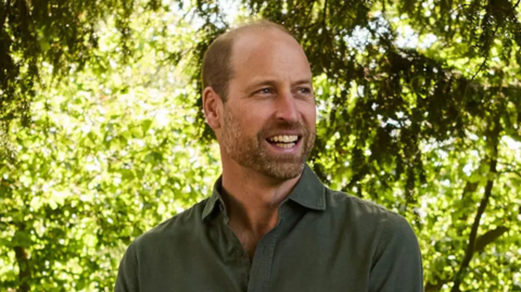 Prince William, standing by trees, wearing a dark green shirt