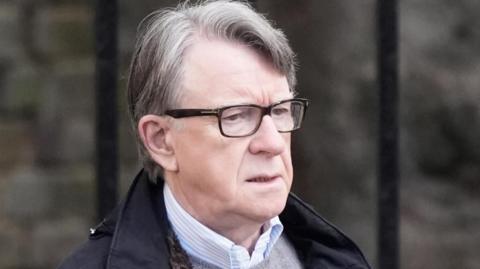 Lord Mandelson wearing glasses, a dark coloured coat, a grey jumper and a shirt.