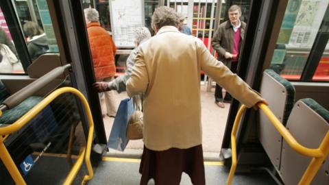An elderly lady in a light mustard coat steps off a bus. She holds on to yellow rails with her right arm. A bus stop with a number of people can be seen in the background.