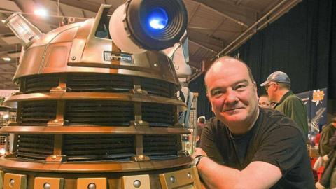 A bronze Dalek with a lit-up blue eye is next to a proud Mike Tucker, who is wearing a black T-shirt, at a convention.