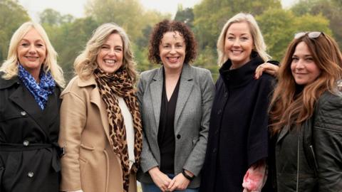 Five women, wearing winter jackets and scarves, standing together in a park on an autumn day. All five are smiling at the camera.