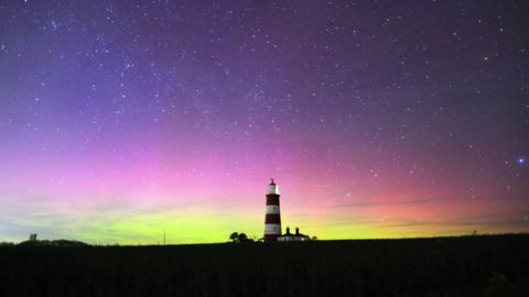 A night-time landscape which has a striped lighthouse is in the middle. The Northern Lights have made the sky turn purple, pink, and yellow, and there are several stars.