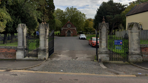 The entrance to a graveyard. The picture is taken from the road and shows the entrance for vehicles with two pedestrian gates either side