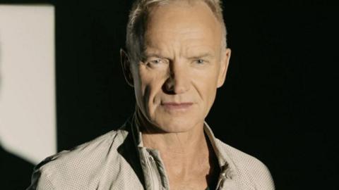 Sting in a casual beige jacket looking thoughtful. His is standing against a black background and to the left part of the wall is lit up. 