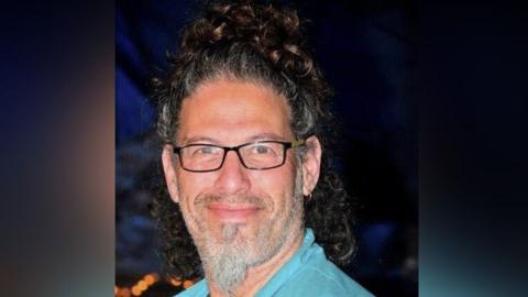 Image shows Ronen Engel in a turquoise blue shirt, with a beard and black glasses smiling with a topknot of dark brown curls on top of his head