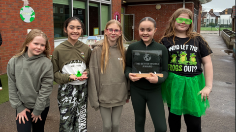 Five primary schoolgirls in Year Six smile at the camera outside a school building. One is wearing a black t-shirt with the logo 'Who let the frogs out', a green tutu and green novelty glasses.
